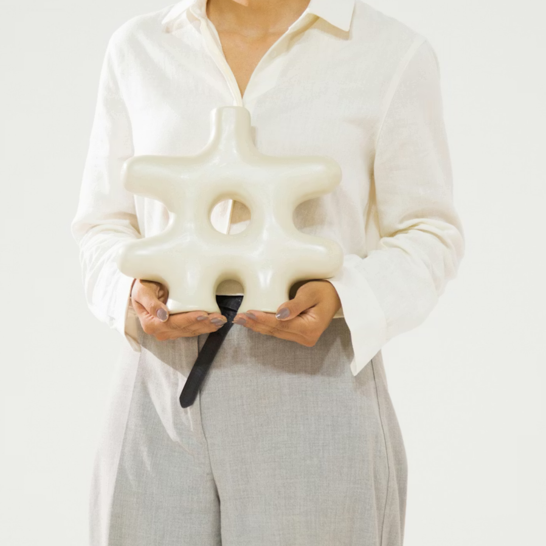 A person in a white shirt and light gray pants holds the Nedjma Vase by Schwung Design—a large, cream-colored handcrafted ceramic piece with rounded, interconnected shapes and a central hole—against a plain background.
