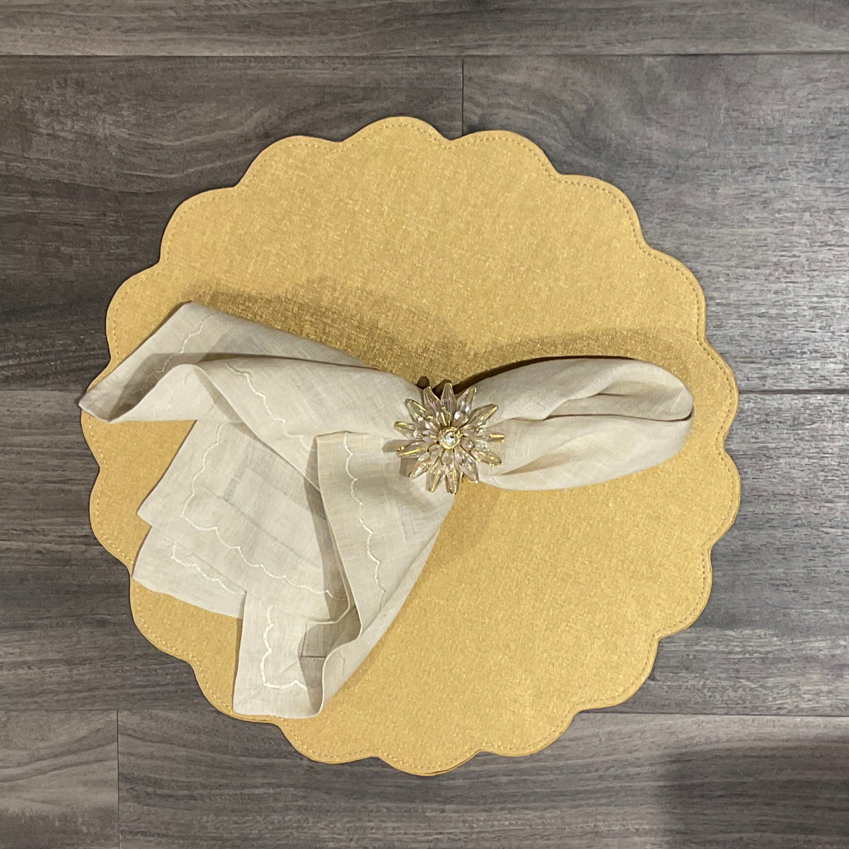 A beige napkin with an embroidered edge, held by a flower-shaped ring, rests on a Victoria Round Scallop Placemat from Deborah Rhodes, arranged on a wood floor.
