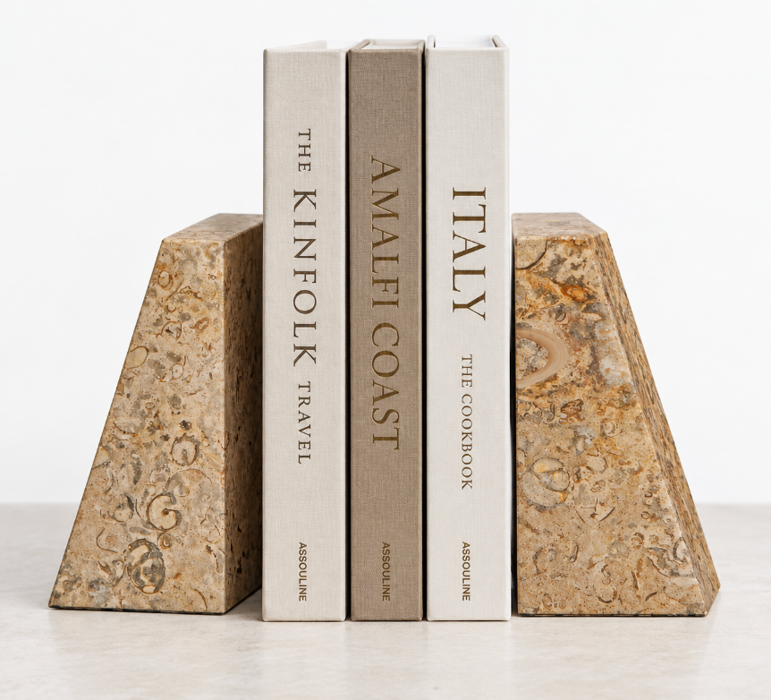 Three books with neutral covers—THE KINFOLK TRAVEL, AMALFI COAST, and ITALY—stand upright between the Zeus Bookend Set in fossil stone by Designs by Marble Crafter on a light surface.