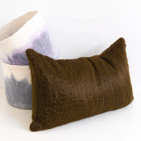 A Woolly Mammoth Pillow in Tobacco by Dove & Donkey (14 x 23) sits in front of two stacked felt baskets, one with a purple and white tie-dye pattern.