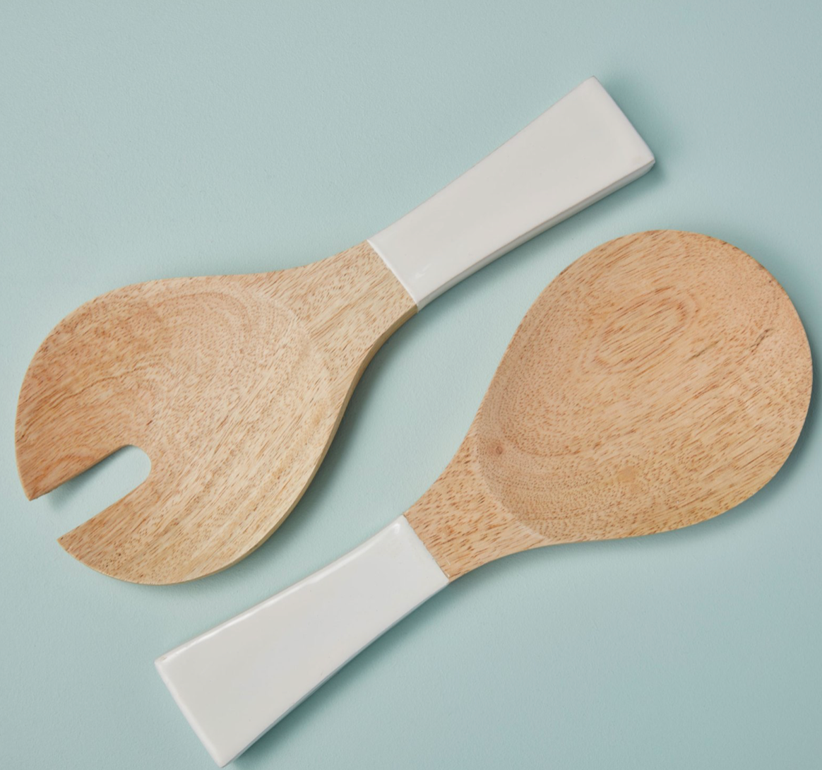 The Mayan Wood Short Salad Servers by Be Home feature white ceramic handles and include a solid spoon and a forked utensil, both handcrafted, placed side by side on a light blue surface.