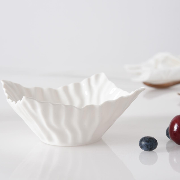 The Mascali Snack Bowl Square by PAMPA BAY, crafted from white porcelain with a textured rim, rests on a glossy white surface. A blueberry and cherry are in the foreground, while the softly blurred background adds a minimal touch.