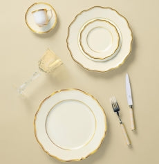 A formal place setting featuring Raynaud's Mazurka Gold & Ivory: a teacup, saucer, three nested Limoges porcelain plates with gold rims, wine glass, and cutlery on beige, showcasing exquisite French craftsmanship.