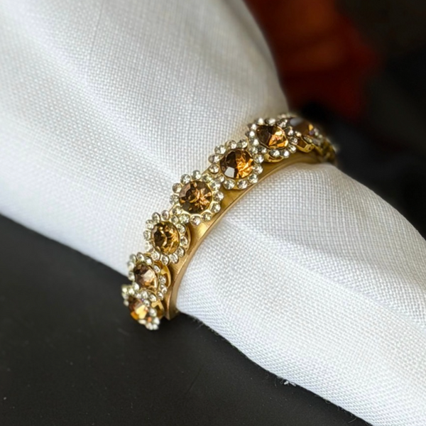 A Golden Bar Napkin Ring set of 4 featuring a gold-toned band with a row of amber crystals surrounded by clear stones, encircling a white cloth napkin.
