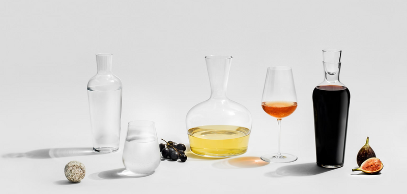 A row of glass bottles and glasses, including the Richard Brendon Jancis Robinson Universal Wine Glass Set of 2, filled with clear, yellow, and amber liquids sits beside grapes, a fig, and cheese on a light surface with a plain background.