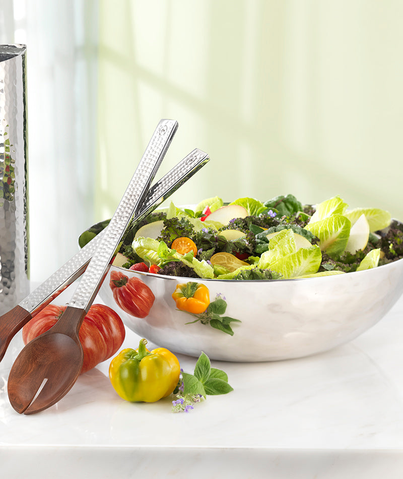 The Mary Jurek Luna Serving Bowl 12" is filled with leafy green salad and vegetables, placed on a marble surface beside wooden salad tongs, fresh tomatoes, and herbs.