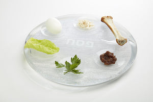 The Apeloig Acrylic Seder Plate elegantly displays a boiled egg, horseradish, roasted bone, lettuce leaf, parsley, and charoset on a clear plate with Hebrew writing at the center.