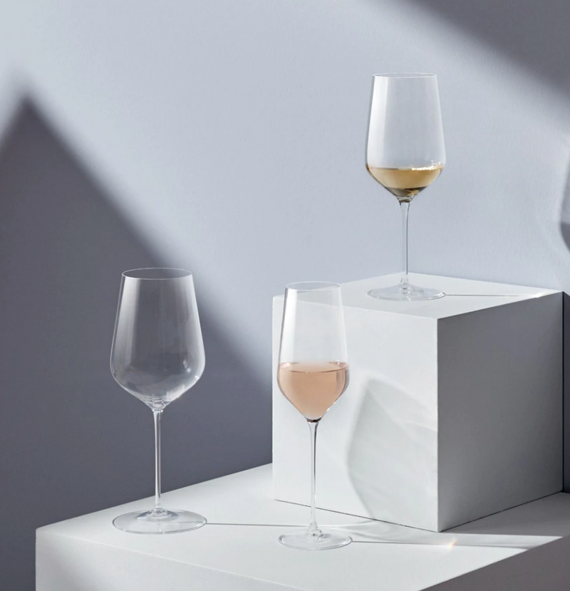 Three Stem Zero Barware wine glasses by Nude Glassware, made from handmade lead-free crystal, are set on white geometric blocks—one empty, one with rosé, and one with white wine—under soft natural light against a pale gray backdrop.