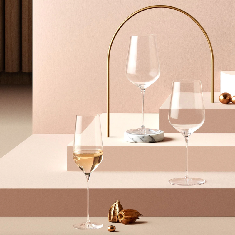 Three elegant Nude Glassware Stem Zero Barware wine glasses, one filled with white wine, are displayed on pastel steps with gold and marble accents. Decorative nuts and a gold arch enhance the minimalist, modern setting.