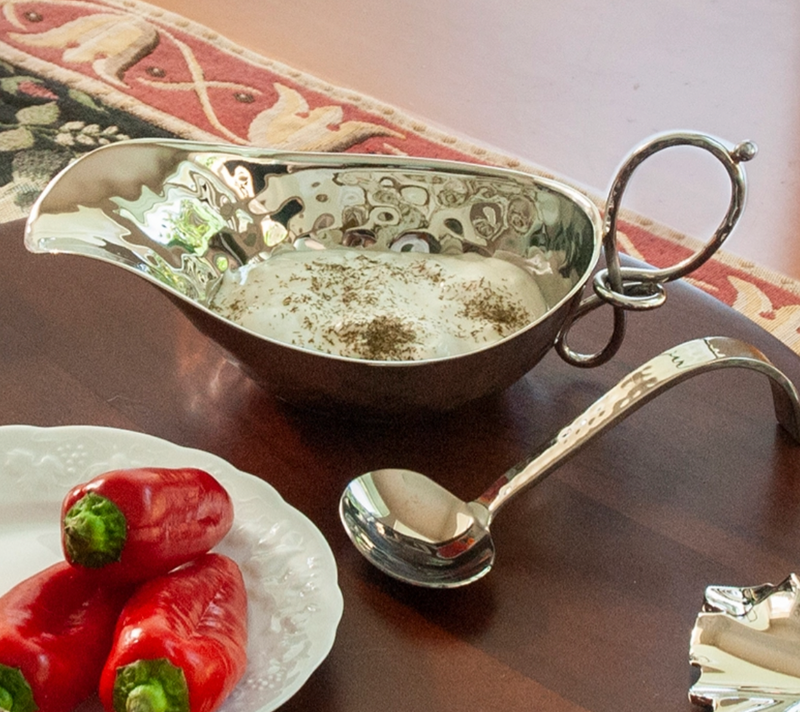 The Mary Jurek Opus Gravy Boat, filled with creamy white sauce and herbs, sits on a table beside red stuffed peppers and a serving spoon.