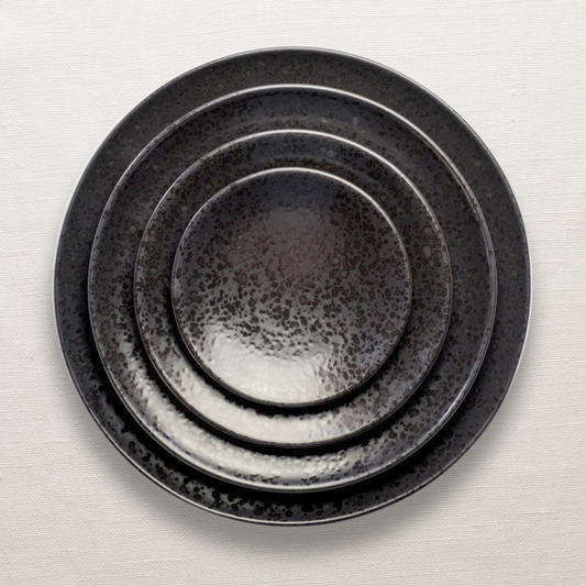 Four round Alchimie Dinnerware Black ceramic plates by L'Objet, featuring a textured speckled finish, are stacked by size on a white tablecloth background.
