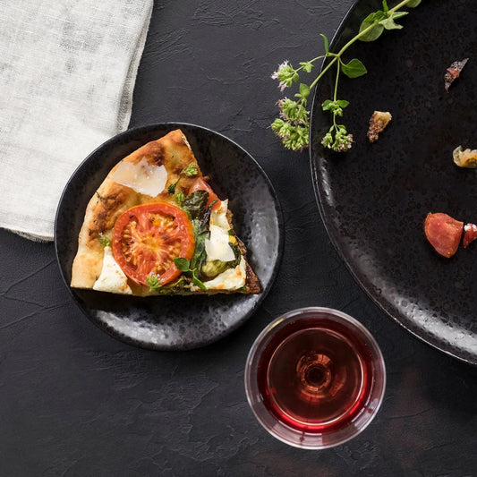 A slice of pizza with tomato, cheese, and basil sits on L'Objet Alchimie Dinnerware Black, next to a glass of red wine, fresh herbs, and an empty plate with crumbs on a dark textured surface.