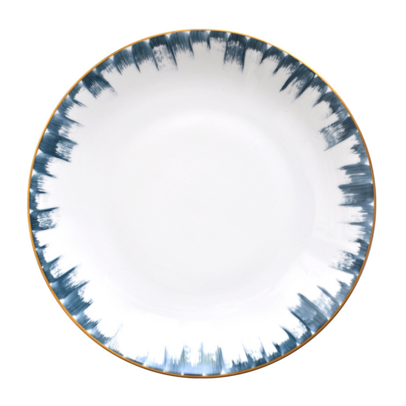 The Marie Daâge Iris 2 Dinnerware features a round white ceramic plate with a gold rim and blue brushstroke pattern, inspired by Marie Daâge’s classic designs. The plain white center enhances its elegant, luxury appeal.