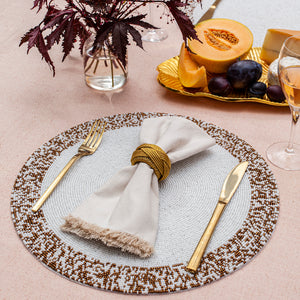 The Splatter Beaded Placemat Set of 4 by Von Gern Home displays a gold fork, knife, and fringed beige napkin in a gold ring on a pink tablecloth, with glasses, dark-leaf vase, and melon and grapes nearby.